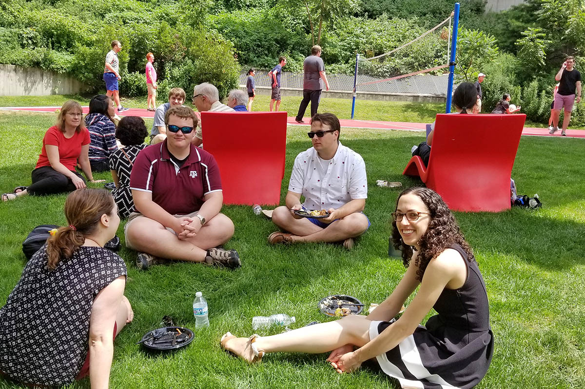 some students sitting on ground eating lunch, some playing volleyball at summer picnic