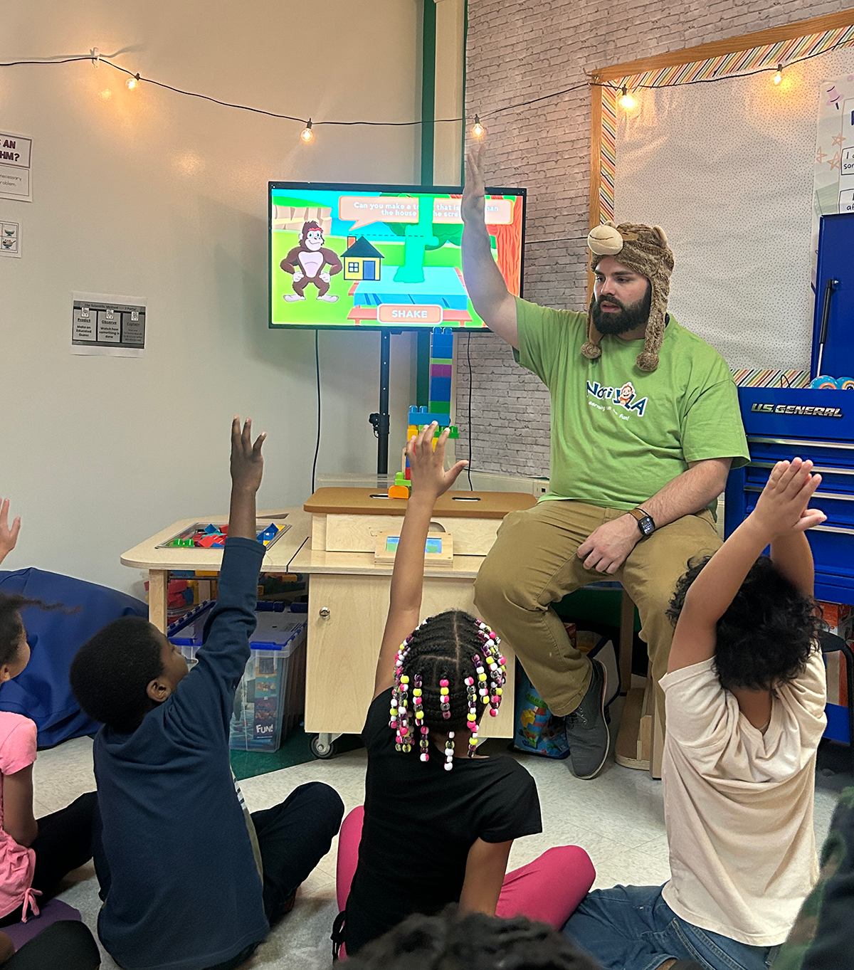 A teacher wears a gorilla hat in the classroom while sitting in front of the Norilla science station. Students are seated on the floor in front of the teacher with their hands raised.