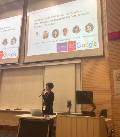 a student presents a research paper at DIS, the title on the large screen behind her reads "Surfacing Design Tensions and Opportunities for AI-Mediated Pre-diagnostic Risk Communication for Breast Cancer Care"