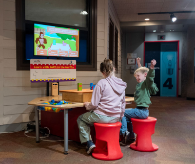 two children seated at the NoRILLA science table, the one raises his hands in the air after getting an answer correct