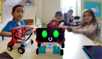 4 children seated around a table, faces blurred for privacy. A small robot sits on the table. Its cartoon face smiles on a digital screen.