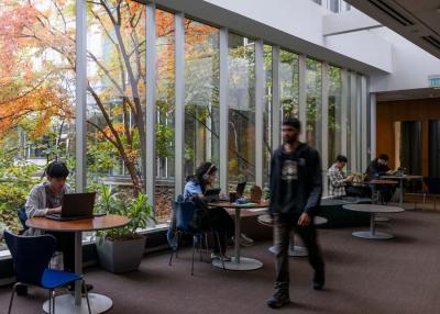 A blurred student walks through a public area where students are working at tables in front of large windows.
