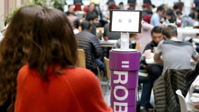 a robot with a purple torso is in the forground of a crowded cafeteria