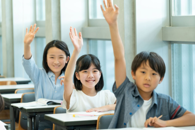 a row of three young students seated at their classroom desks, all 3 have their hand raised