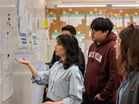 Team members are huddled together and face a wall covered in wireframes. There is a large affinity diagram on the far wall behind them.