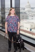 Chancey stands in front of a large window with many buildings in the distance, a black lab service dog sits at her side