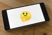 A mobile phone laying on a wooden table top. The screen is white with a yellow "thinking face" emoji in the center.