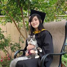 Connie is wearing her black CMU cap and gown. She is sitting outdoors in a chair and holding her cat.