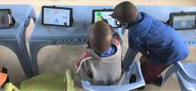 the view of the back two young boys interacting with a touchscreen tablet sitting on a plastic table