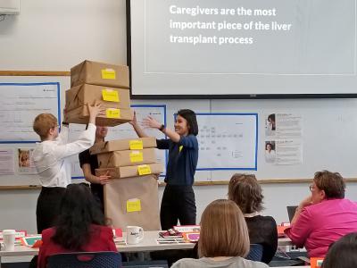 students stack a pile of boxes in the arms of a teammate to illustate the burden of being a caregiver during a capstone presentation