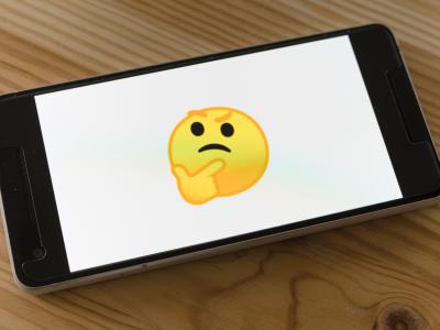 Mobile phone on wooden table. The screen is white with a yellow "thinking face" emoji in the center.