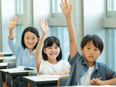 a row of three young students seated at their classroom desks, all 3 have their hand raised