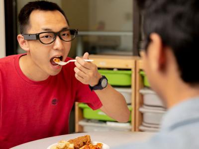 A person sitting at table eating bite of food. There is a small sensor on the side of their glasses