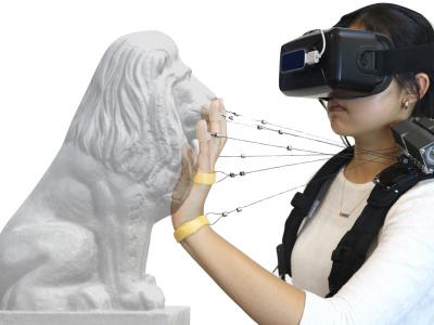 student wearing VR headset on face touches the face of a lion statue while wires connect fingers to the Wireality device on her shoulder