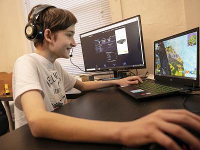 young student smiles while sitting at desk with laptop and extra screen and playing a game online