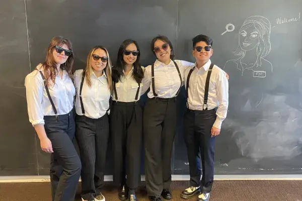 the 5 student team stands side by side, all wearing matching black pants, white shirts, black suspenders, and sunglasses