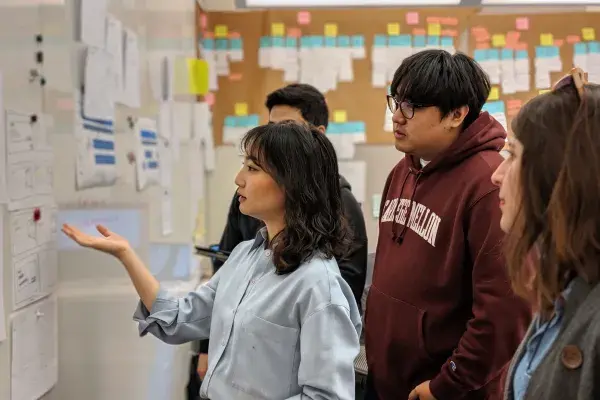 Team members are huddled together and face a wall covered in wireframes. There is a large affinity diagram on the far wall behind them.