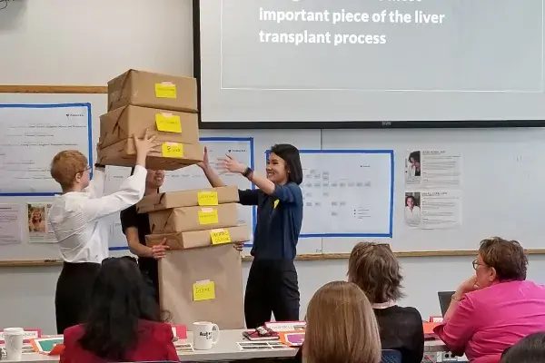 students stack a pile of boxes in the arms of a teammate to illustate the burden of being a caregiver during a capstone presentation