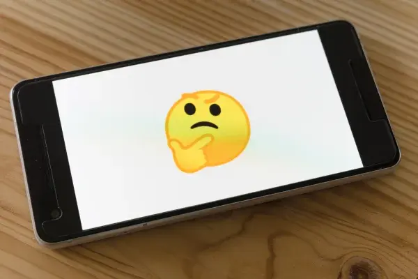 Mobile phone on wooden table. The screen is white with a yellow "thinking face" emoji in the center.