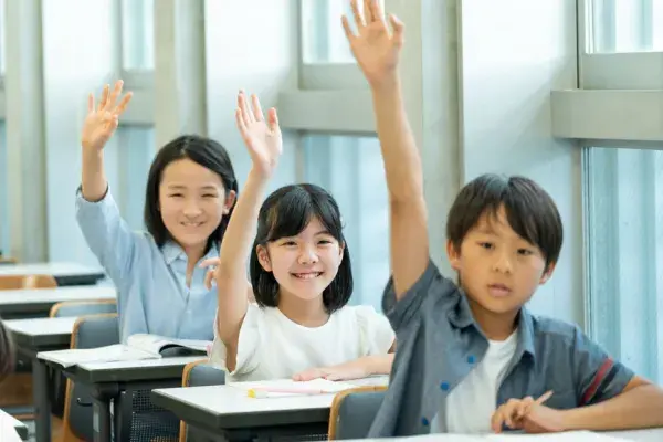 a row of three young students seated at their classroom desks, all 3 have their hand raised