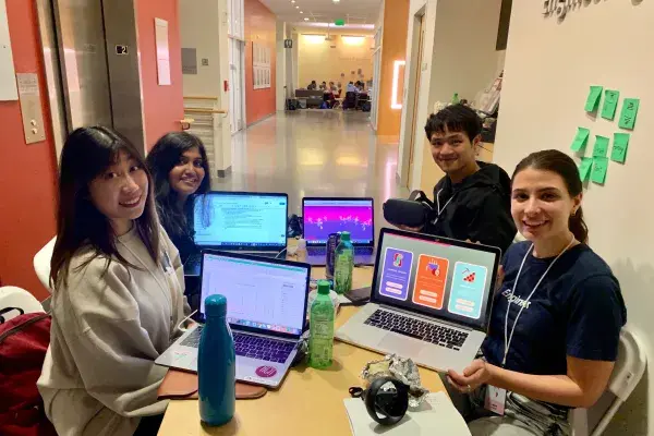 4 students at a table during the hackathon & show their laptop screens while working on Lucid Drums