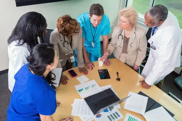 6 doctors and nurses stand around the end of a table looking at charts, graphs and a laptop