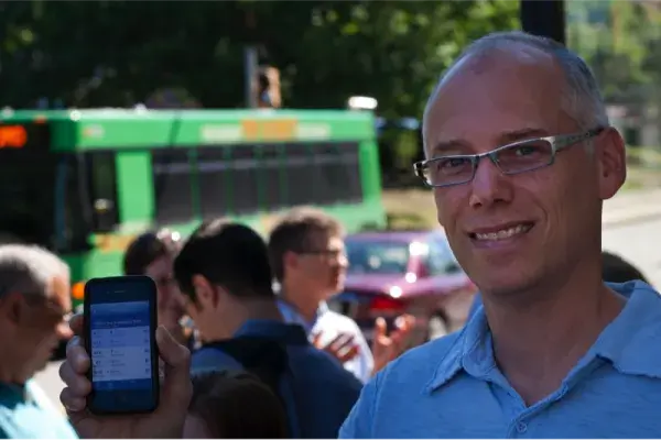 John Zimmerman smiles and stands at a busy bus stop. He's holding up a mobile phone with the Tiramisu app visible on the screen