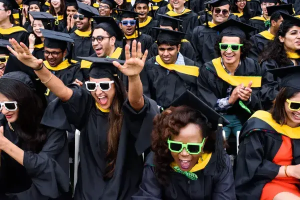 A close up of the crowd celebrating during commencement. These SCS grads are wearing black robes with a yellow hood and also wearing matching 8bit sunglases